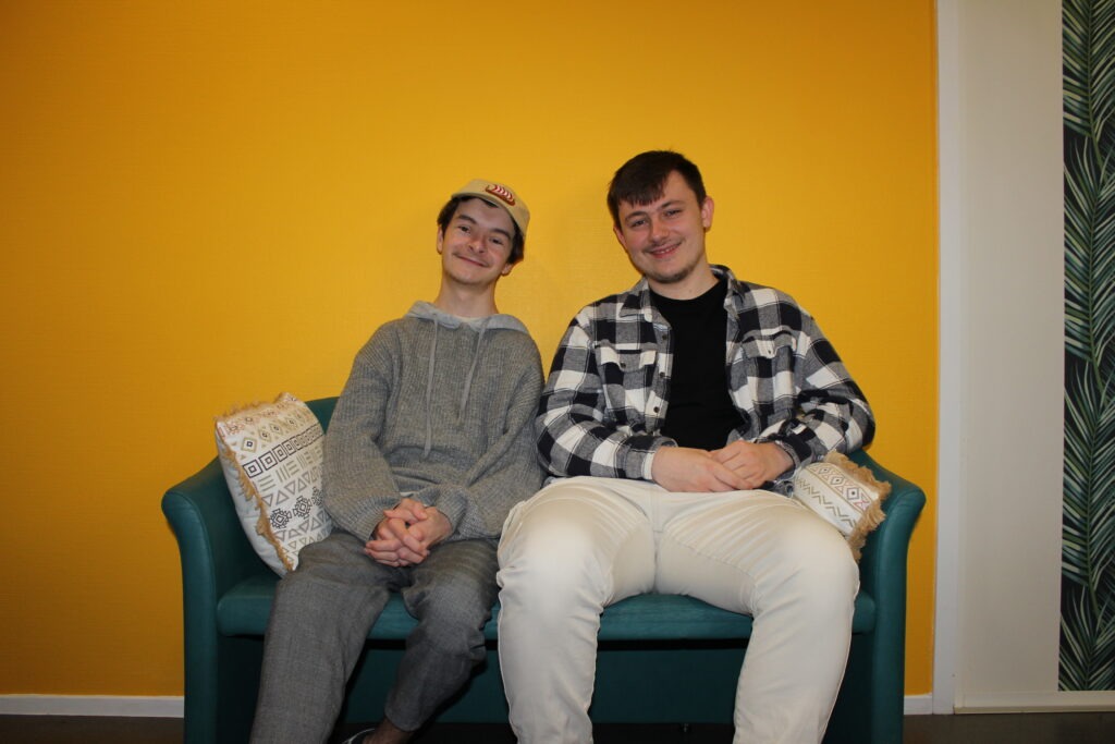 Deux jeunes souriants assis sur un canapé vert, sur un fond jaune.